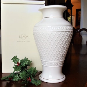 May include: A cream-colored Lenox American by Design vase featuring a textured diamond lattice pattern, embossed scrollwork borders, and gold-tone rim accents, accompanied by a sprig of artificial holly with red berries.