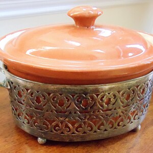 May include: A terracotta-colored ceramic casserole dish with a matching lid and a silver-colored metal filigree stand. The dish has a round shape and small handles. The stand has decorative cutouts and small feet.