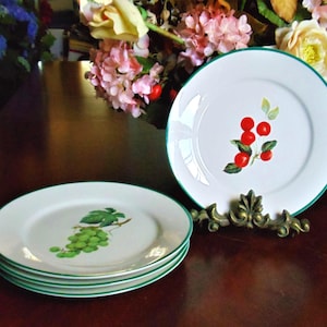 May include: White ceramic plates with a green trim and fruit designs. One plate features a cluster of red cherries with green leaves, while others display green grapes. The plates are stacked and displayed on a dark wooden surface.