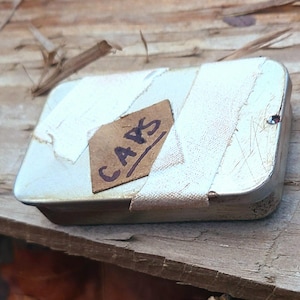 以下が含まれることがあります： 白い塗装が施された小さな長方形の金属製の箱で、「CAPS」と書かれた茶色のラベルが付いています。 箱は白い布で包まれ、木製の表面に置かれています。 この箱はおそらく保管用に使用されています。