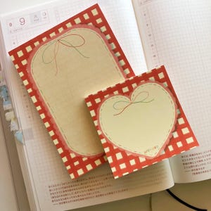 Peut inclure: Deux blocs-notes décoratifs avec un motif à carreaux rouges et blancs. L'un est rectangulaire, l'autre en forme de cœur. Les deux ont un centre crème avec une bordure cousue et un nœud.