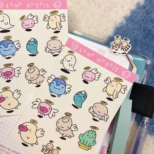May include: Two sheets of stickers featuring various cartoon characters with halos and wings. The stickers are on a white background and have a pink border. The text "tama angels" is written on the top of each sheet.