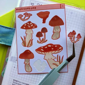 May include: A sheet of stickers featuring various red and white mushrooms with white spots. The stickers are on a white background with a pink title bar that says "mushrooms.png".