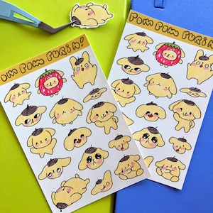 May include: Two sticker sheets featuring yellow cartoon characters with brown berets and various expressions, along with a red strawberry character. The text "Pom Pom Purin!" is printed at the top of each sheet. A single sticker and tweezers are also visible.