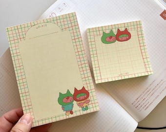 May include: Two sets of sticky notes with a grid pattern. One set has a larger note with a red, green, and yellow border and a smaller note with a red, green, and yellow border. Both sets feature a cartoon illustration of a cat with a red and green hat. The larger note has the word "WORK" written on it. The smaller note has the word "WORK" written on it.