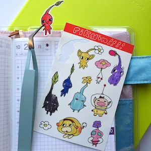 May include: A sheet of stickers featuring various cartoon characters, including a purple creature with a long neck, a yellow creature with a flower on its head, and a blue creature with a flower on its head. The stickers are on a white background and have a pink border. The text "PIKAOU!!" is written in red at the top of the sheet.