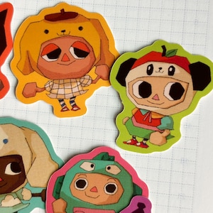 May include: A collection of colorful die-cut stickers featuring cartoon characters in various costumes. The stickers include characters dressed as a dog, an apple, and a dinosaur. The stickers are on a white grid background.