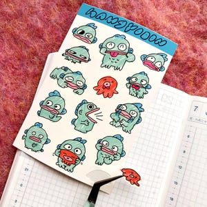 Op de afbeelding: Een vel stickers met cartoonvissen en een octopus. De vissen zijn lichtblauw met roze accenten en verschillende uitdrukkingen. Het woord "HANGYOON" staat in blauw bovenaan het vel. Een pincet houdt een kleine octopussticker vast.