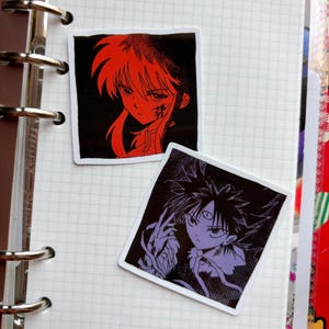 Puede incluir: Dos pegatinas cuadradas con ilustraciones de estilo anime. Una pegatina tiene un personaje rojo sobre fondo negro. La otra pegatina tiene un personaje morado sobre fondo negro. Ambas pegatinas est&aacute;n sobre un cuaderno de papel cuadriculado.