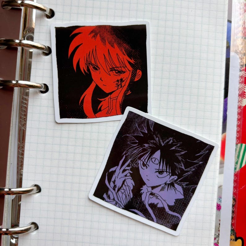 Puede incluir: Dos pegatinas cuadradas con ilustraciones de estilo anime. Una pegatina tiene un personaje rojo sobre fondo negro. La otra pegatina tiene un personaje morado sobre fondo negro. Ambas pegatinas est&aacute;n sobre un cuaderno de papel cuadriculado.