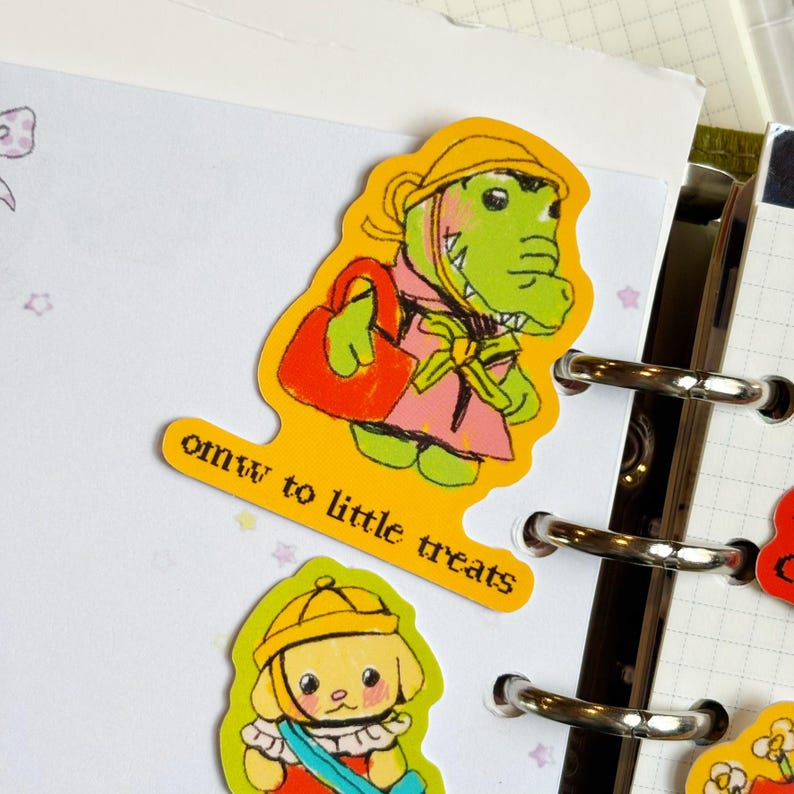 May include: A yellow sticker featuring a cartoon alligator wearing a pink coat and yellow hat, carrying a red purse. The sticker has the text "omw to little treats". The sticker is placed on a white paper with a planner.