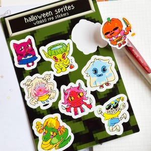 May include: A sheet of Halloween-themed stickers featuring colorful cartoon sprites. The stickers include a pink cat, a green-skinned creature, a blue ghost, a red devil, and a skeleton. The sheet has the text "halloween sprites" and "w0nkko rpg stickers".