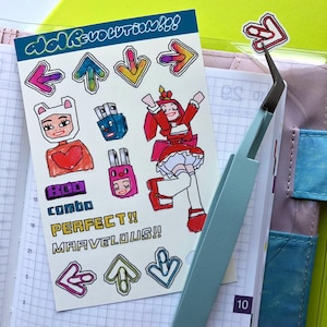 May include: A sheet of stickers featuring a cartoon character in a red dress, a white bear with a pink heart, and various arrows. The text "combo", "PERFECT!!", and "MARVELOUS!!" is also included on the sheet.