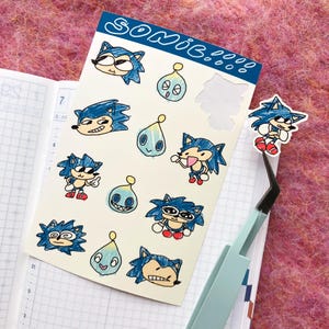 May include: A sheet of stickers featuring various cartoon characters from the Sonic the Hedgehog franchise. The stickers are drawn in a simple, hand-drawn style and feature characters like Sonic, Tails, and Knuckles. The stickers are on a white background with the word "SONIC!" written in blue at the top.