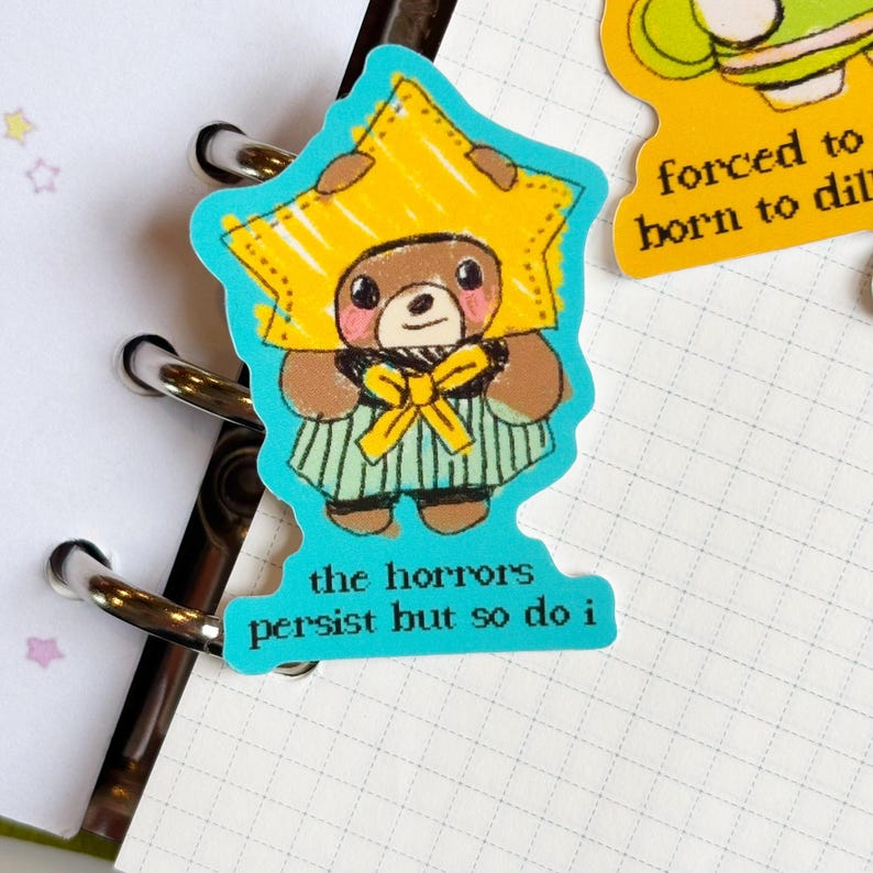 May include: A die-cut sticker featuring a cartoon bear wearing a yellow star-shaped hat and a green and white striped outfit. The sticker has the text "the horrors persist but so do i".