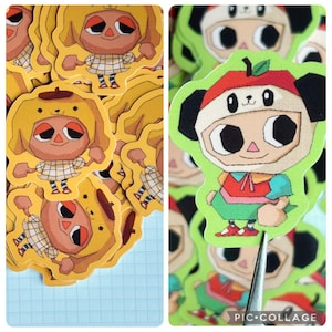 May include: A collection of die-cut stickers featuring cartoon characters. One design shows a character in a yellow dog costume, and the other shows a character in a panda costume with an apple on its head. The stickers have a white border.