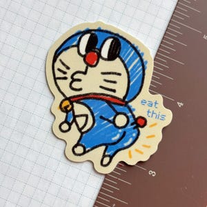 Op de afbeelding: Een gestanste sticker met een blauw stripfiguur met een rode kraag en een bel. Het personage blaast een kus, met de tekst "eat this" in handschrift. De sticker heeft een gele rand.