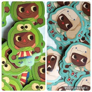 May include: A collection of colorful stickers featuring cartoon characters. One design shows a character in a green frog costume, while another depicts a character in a white hooded outfit. The stickers have a whimsical, illustrated style.