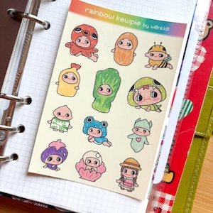 May include: A sheet of colorful stickers featuring cartoon characters dressed as various foods and creatures. The stickers include an octopus, peanut, bee, and frog. The text "rainbow kewpie by wønkkø" is printed at the top.