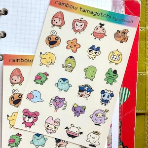 May include: Two sheets of colorful stickers featuring various cartoon characters. The stickers are labeled "rainbow tamagotchi by w0nkko" and "rainbow". The characters are in a variety of colors and designs, including animals and other whimsical figures.