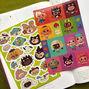 May include: Two sticker sheets featuring colorful cartoon characters. One sheet has a green background with various character designs. The other sheet has a grid of characters on a red, orange, and pink background. The stickers are on a notebook.
