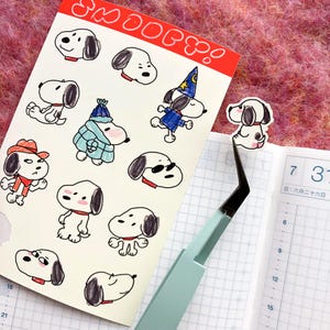 May include: A sheet of Snoopy stickers with various Snoopy illustrations, including Snoopy in a cowboy hat, a wizard costume, and sunglasses. A sticker is held by tweezers next to a planner. The word "Snoopy" is written at the top.