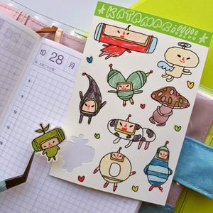 May include: A sheet of colorful stickers featuring cartoon characters from the Katamari Damacy video game series. The stickers are drawn in a simple, childlike style and feature a variety of characters, including a mushroom, a cow, and a pencil.