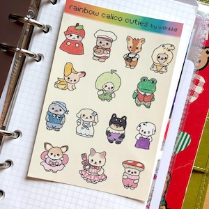 May include: A sheet of stickers featuring colorful cartoon characters. The stickers include various animals and food-themed characters, such as a strawberry, a chef, and a frog. The text "rainbow calico cuties by w0nkko" is printed at the top.