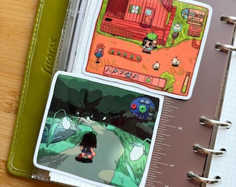Könnte beinhalten: Zwei quadratische Aufkleber mit Szenen aus einem Videospiel. Einer zeigt eine Figur vor einem roten Haus mit Garten und Hühnern. Der andere zeigt eine Figur, die auf einem Weg in einem dunklen Wald geht. Die Aufkleber befinden sich auf einem Planer mit einem Lineal.