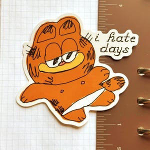 以下が含まれることがあります： 「i hate days」の文字が入ったオレンジ色のガーフィールドのステッカー。不機嫌そうな表情の漫画のガーフィールドが描かれています。ステッカーは白い背景にあります。