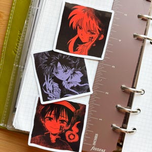 Puede incluir: Tres pegatinas cuadradas con ilustraciones de personajes de estilo anime. Una pegatina presenta un personaje de tonos rojos con pelo largo, otra un personaje de tonos morados con tres ojos y la tercera un personaje de tonos rojos con un sombrero.