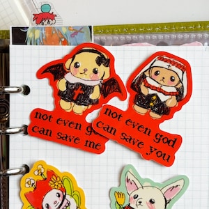 May include: Red stickers with cartoon dog illustrations. One dog is dressed as a devil, the other as a nun. Text on the stickers reads "not even god can save me" and "not even god can save you". The stickers are on a white grid paper.