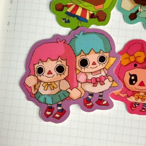 May include: A collection of colorful stickers featuring cartoon characters. One sticker shows two figures with pink and blue hair, holding hands. They wear dresses and have large, round eyes. Other stickers show similar characters in various outfits.