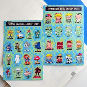 May include: Two sticker sheets with colorful cartoon characters. One sheet is labeled "mother enemies sticker sheet" and the other is labeled "earthbound buds sticker sheet". The stickers are on a light blue background with a grid pattern.