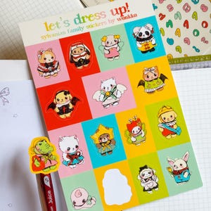 May include: A sheet of colorful Sylvanian Family stickers by wonkko. The stickers feature various animal characters dressed in costumes, set against a grid of pink, red, yellow, and green squares. The text "let's dress up!" is at the top.