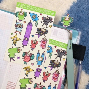 May include: A sheet of colorful stickers featuring cartoon characters with robot-like features. The stickers are on a white background with a green border. The stickers are arranged in a random pattern. The text "KATAMARI DAMO" is at the top of the sheet.