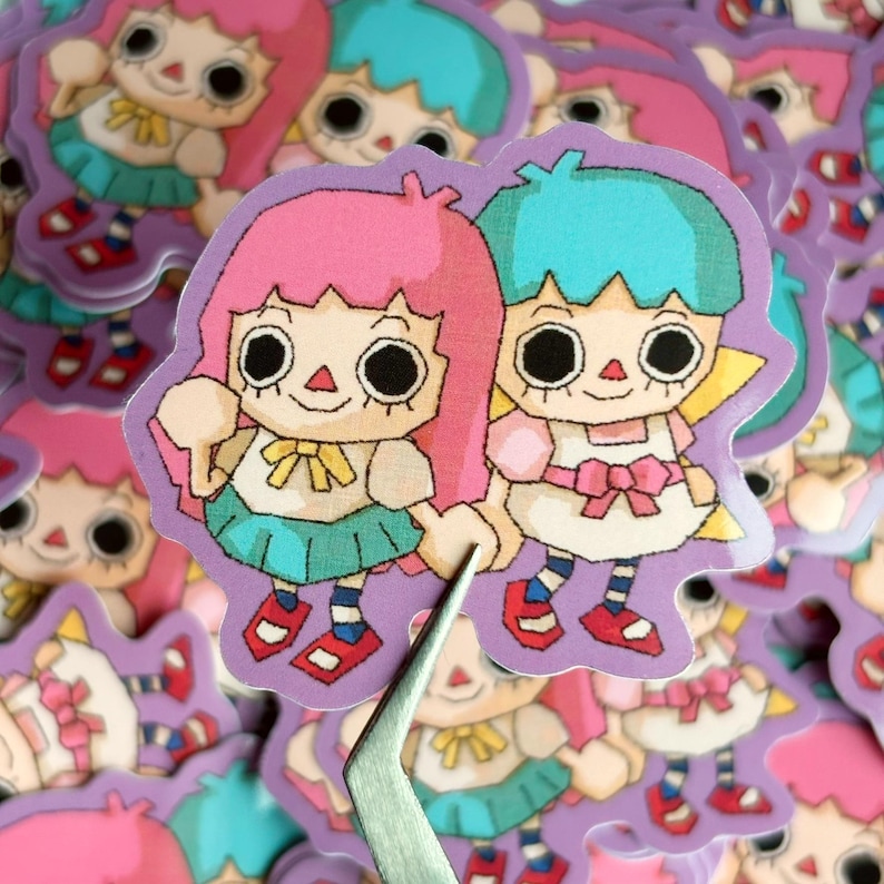 May include: A collection of stickers featuring two cartoon characters with large, black eyes, holding hands. One character has pink hair, the other has blue hair. They wear dresses and are set against a purple background.
