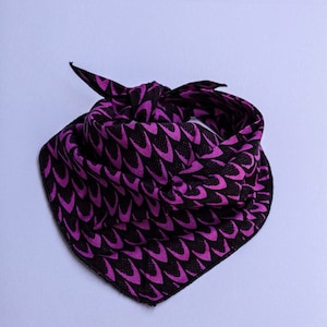 May include: A triangular bandana tied in a knot, featuring a repeating pattern of purple, curved, scale-like shapes set against a solid black background. The fabric is folded to create a layered appearance with visible edges and a pointed bottom.