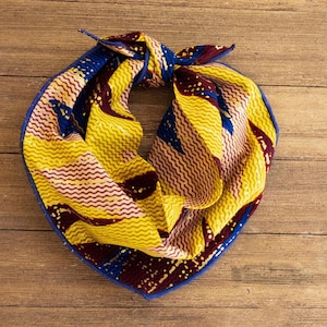 May include: A colorful scarf with a vibrant pattern of yellow, blue, and burgundy zigzags. The scarf is tied in a knot, showcasing its intricate design and contrasting blue border. The scarf is made of a soft fabric.