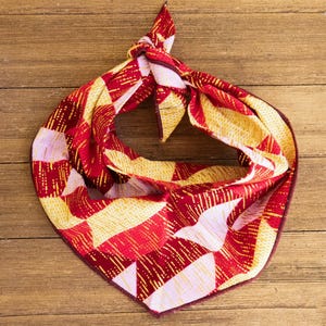 May include: A red, yellow, and white patterned scarf with a knotted design. The scarf features a geometric pattern of triangles and lines. The scarf is made of a soft material and is displayed on a wooden surface.