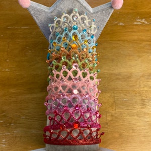 Pode incluir: Uma pilha de coroas de princesa coloridas com glitter e strass em um suporte em forma de coroa cinza.
