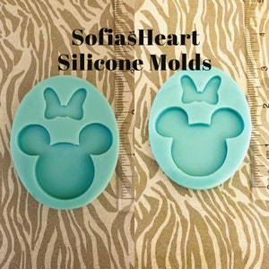 May include: Two light blue silicone molds shaped like a cartoon mouse head with a bow on top. The molds are on a zebra patterned surface. The text "SofiasHeart Silicone Molds" is above the molds.