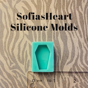 May include: A turquoise silicone mold shaped like a coffin, measuring approximately 1 inch in length. The mold is sitting on a ruler with markings in inches.