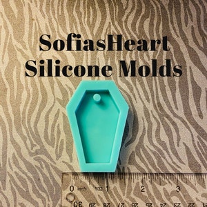 May include: A turquoise blue silicone mold shaped like a coffin with a small hole at the top. The mold is about 1 inch wide and 2 inches long. The mold is sitting on a brown and white zebra patterned surface.