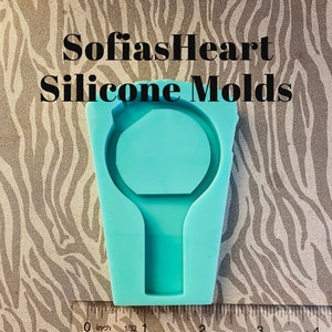 May include: A turquoise silicone mold for making a ring with a round center and a small handle. The mold is about 2 inches in diameter and is shown next to a ruler.