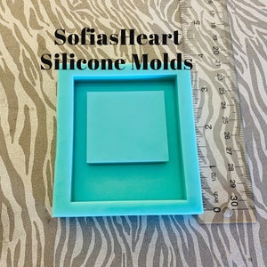 May include: A square, light blue silicone mold with a smaller square inset. The mold is labeled "SofiasHeart Silicone Molds." A ruler is placed next to the mold, showing measurements in inches. The background has a zebra-stripe pattern.