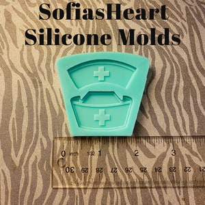 May include: A teal blue silicone mold shaped like a nurse's hat with a white cross on each hat. The mold is about 1 inch wide and is sitting on a ruler with inch markings.