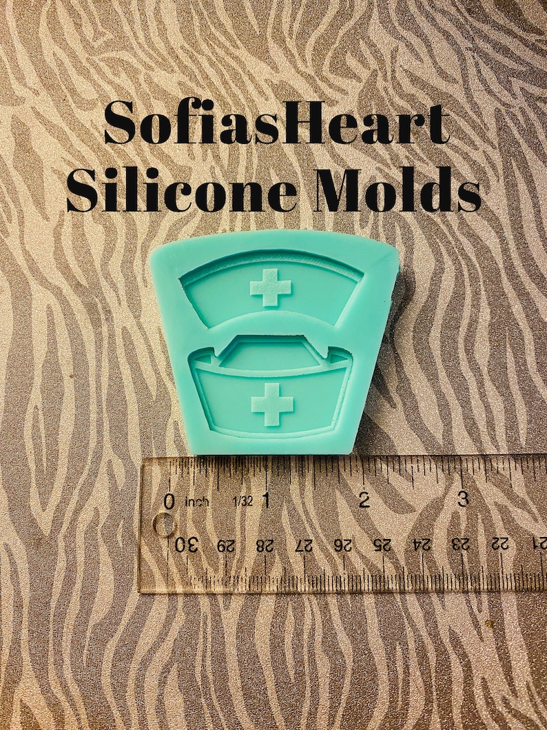 May include: A teal blue silicone mold shaped like a nurse's hat with a white cross on each hat. The mold is about 1 inch wide and is sitting on a ruler with inch markings.