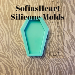 May include: A turquoise blue silicone mold shaped like a coffin. The mold is approximately 6 inches long and 3 inches wide. The mold is on a zebra print background.