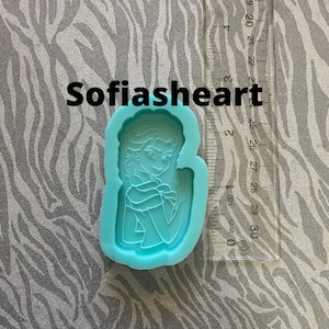May include: A light blue silicone mold of a cartoon character with long blonde hair, wearing a blue dress and a white cape. The mold is on a white and gray patterned surface.
